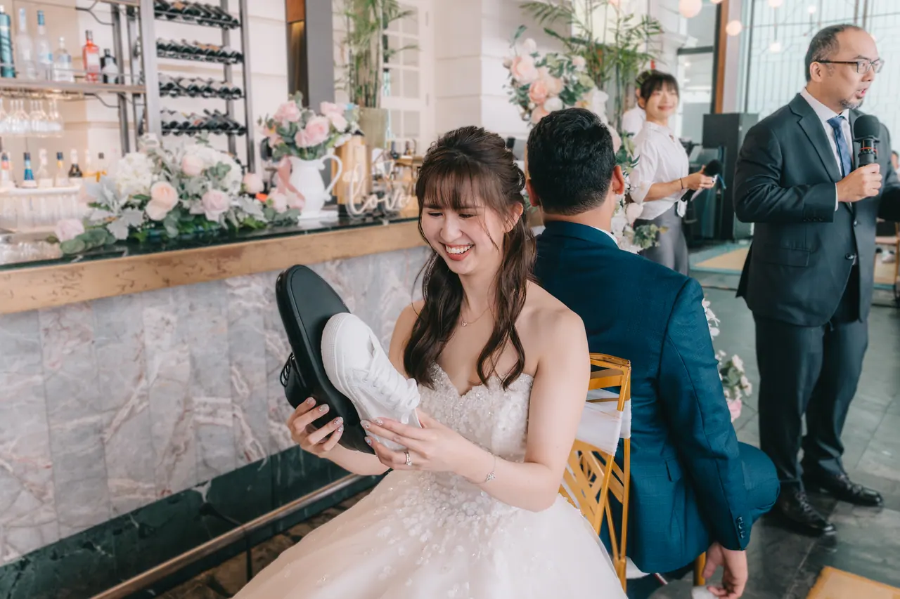 The bride holds up a shoe during a game, guests laughing around her.