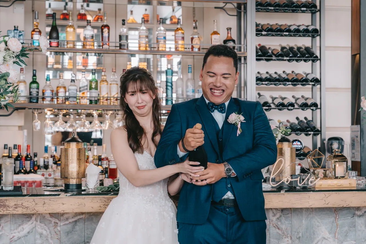 The couple laugh together in front of a wall of bottles.