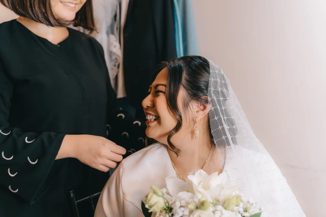 A friend helps the bride into her veil; the bride laughs.
