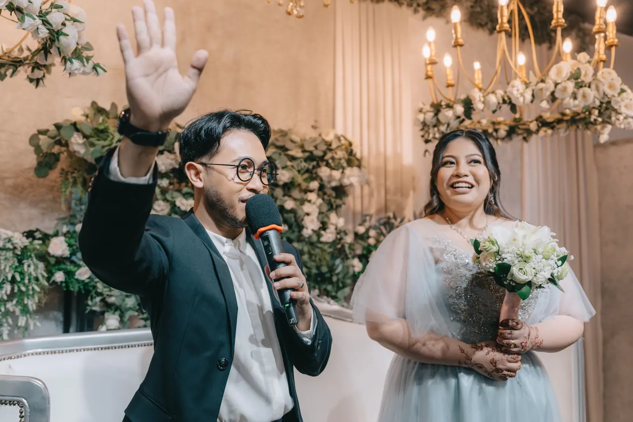 The groom waves mid-speech; the bride laughs beside him.