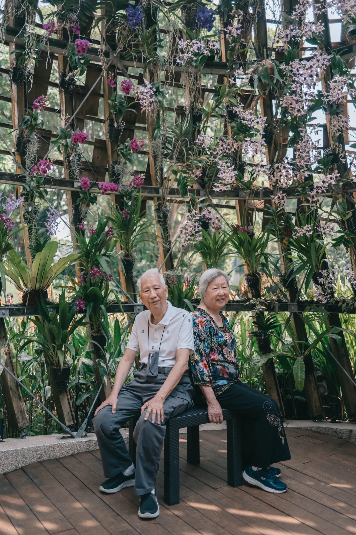 An older couple sit together among hanging orchids.