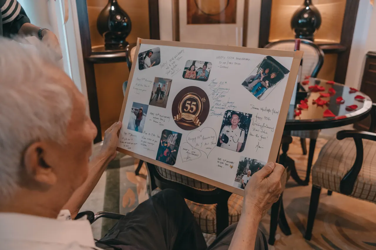An older man holds up an anniversary board covered in photos.