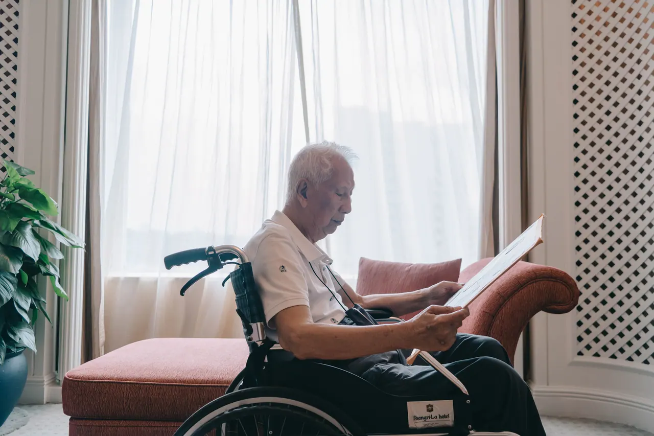 An older man in a wheelchair reads by the window.