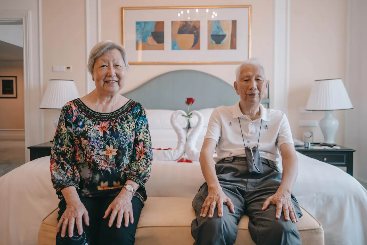 An older couple sit side by side on a hotel bed, smiling.