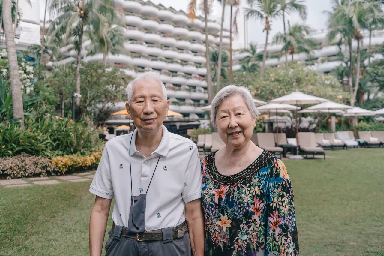 An older couple stand on a lawn in front of a hotel.