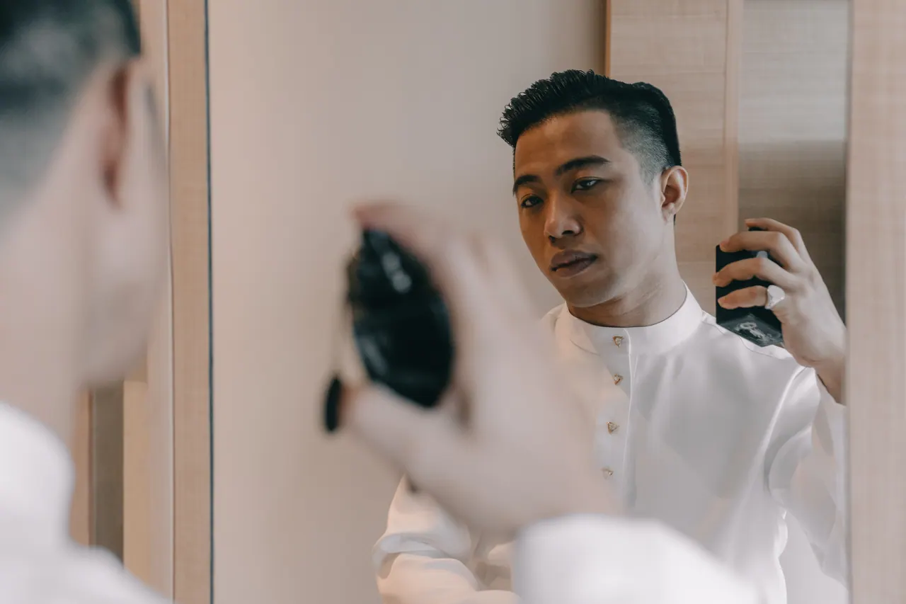 The groom sprays perfume, seen in a mirror.