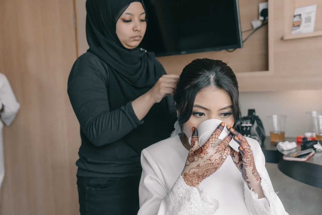 A make-up artist works on the bride as she holds a cup.