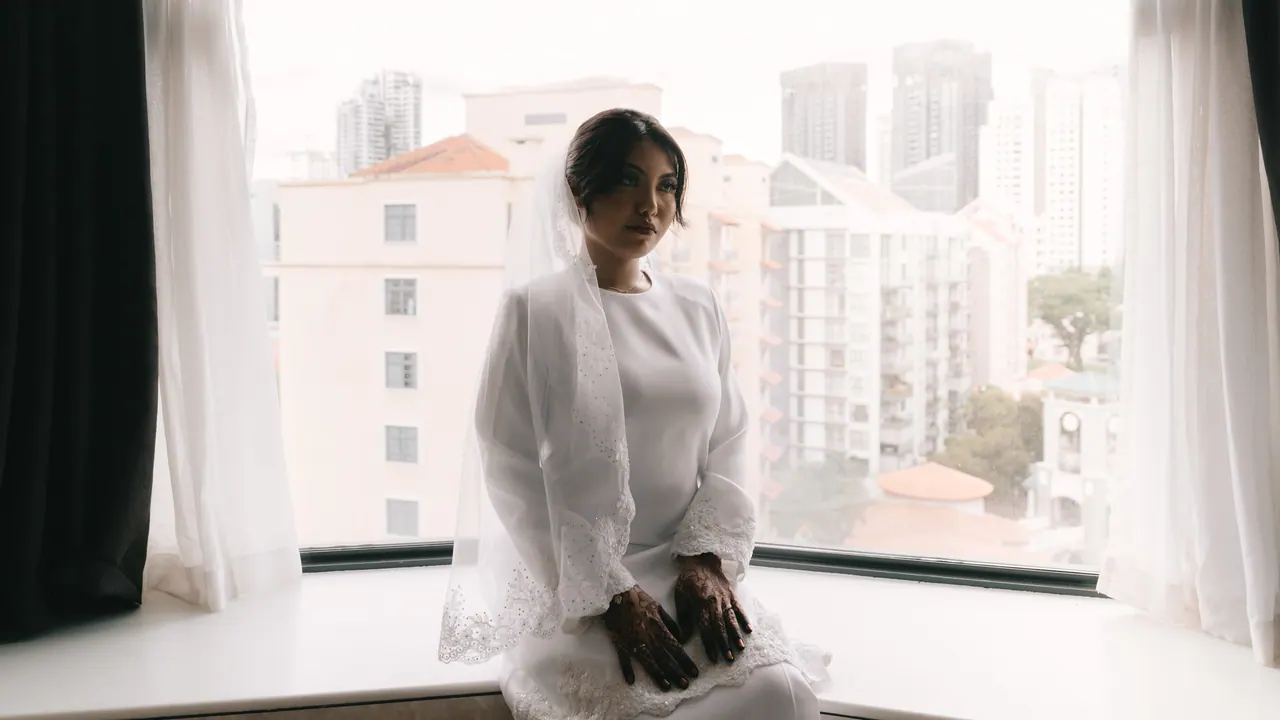 The bride sits by a tall window overlooking the city.
