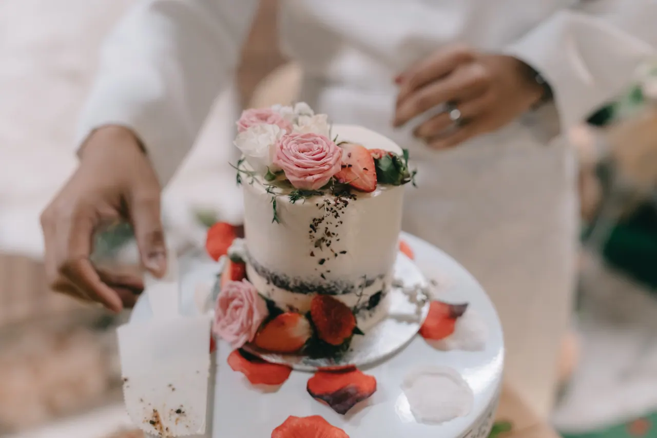 Hands cut a small white cake topped with flowers and strawberries.