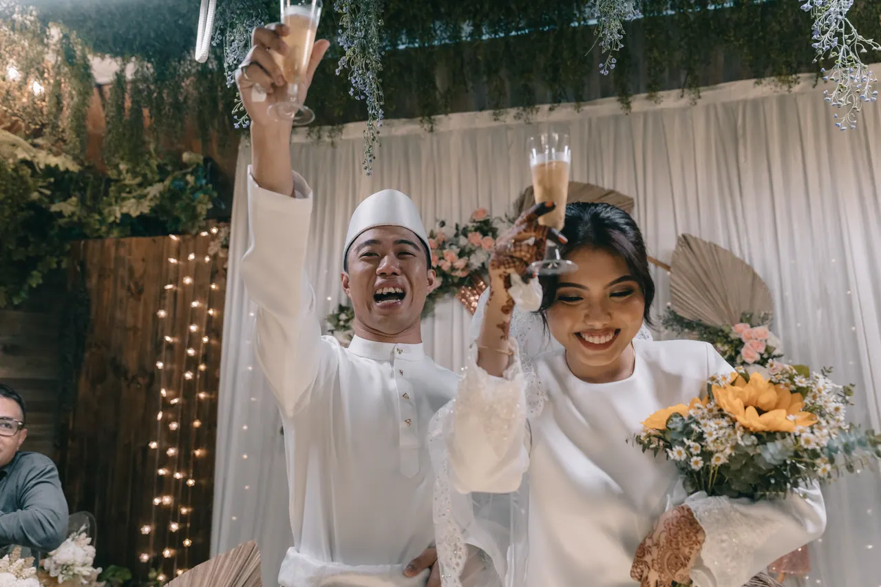 The couple raise their glasses, laughing, in front of the floral arch.