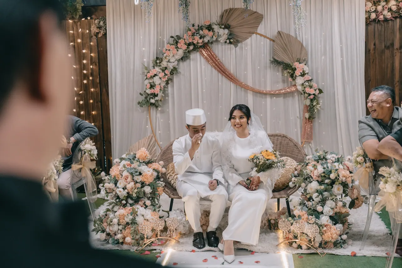 The couple seated on the dais beneath a floral arch.